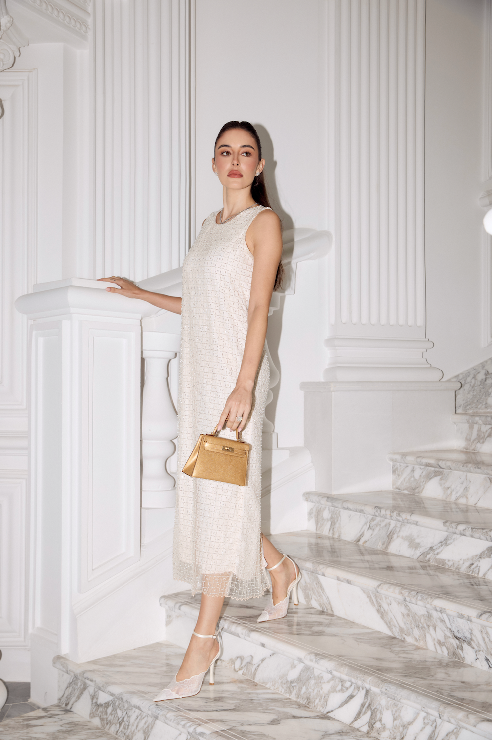 A woman stands on white marble stairs in a bright white interior, wearing a sleeveless textured cream dress and holding a small tan handbag.