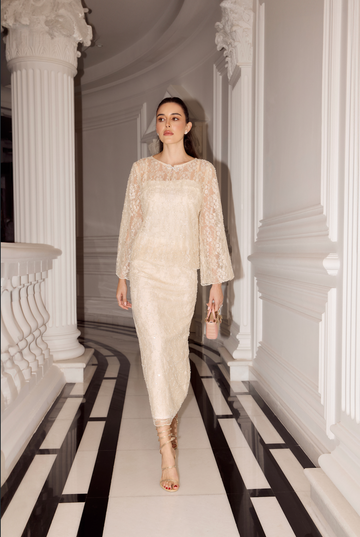 A woman walks down a grand hallway wearing a cream lace and satin midi dress with long sleeves and high neck, paired with nude high-heeled sandals and carrying a small beige handbag.
