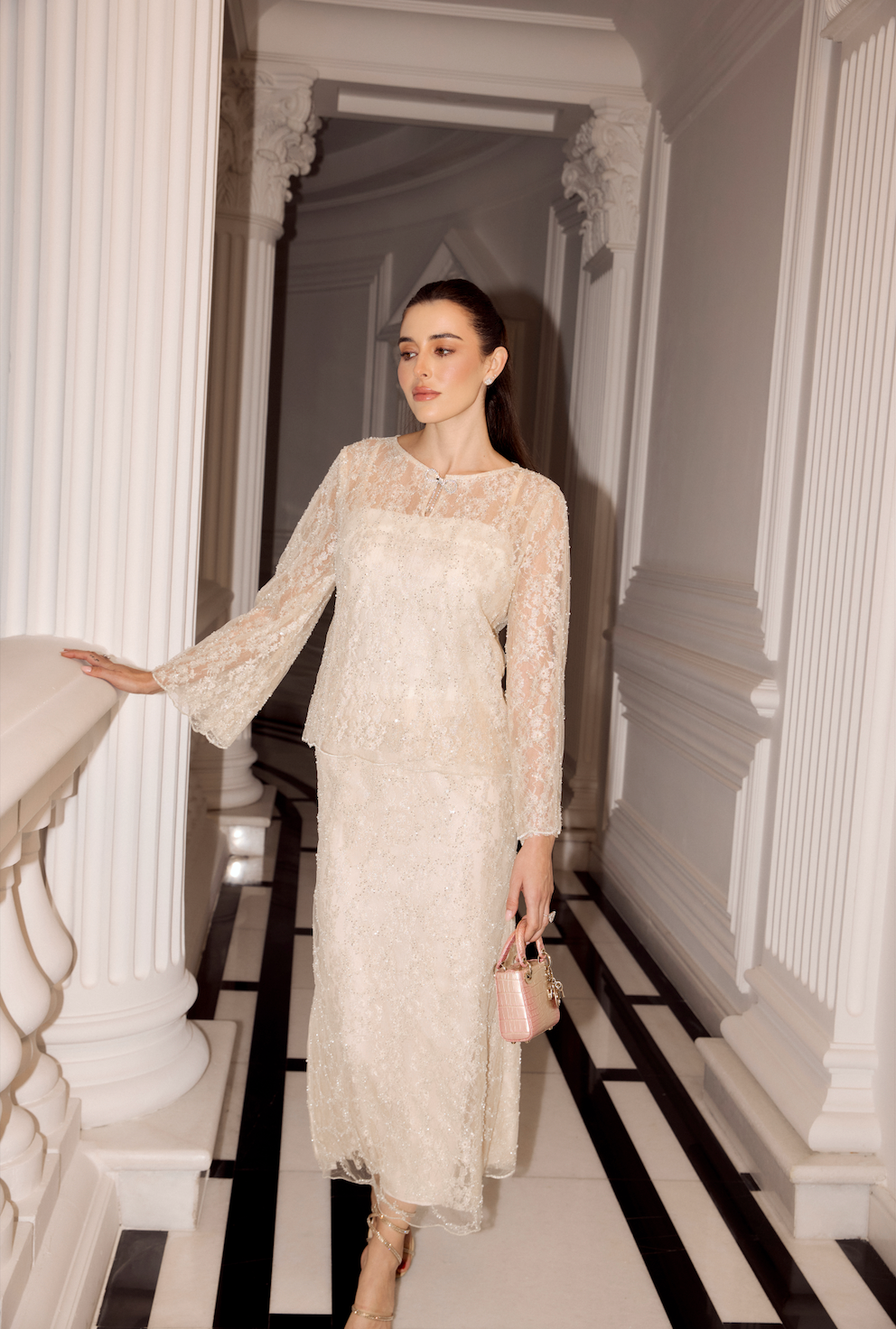 A woman wearing a cream lace midi dress with long sleeves and a sheer yoke walks down a grand white hallway with black-and-white tiled floors.