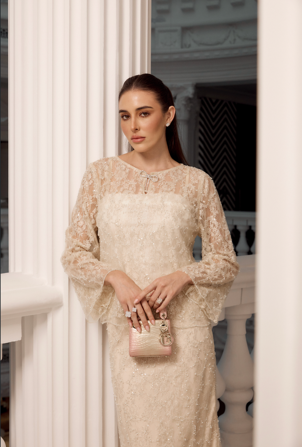 A woman stands indoors wearing a fitted lace and sequin cream dress with long flared sleeves and holding a small pink textured handbag.