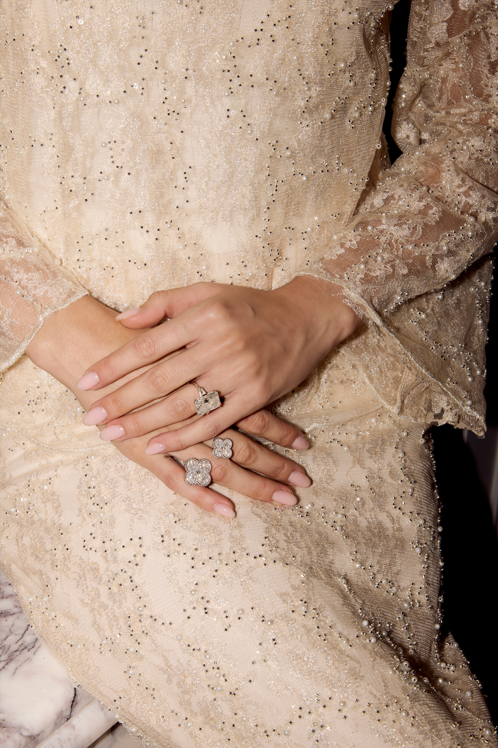 A close-up of a person's hands wearing four large, sculptural silver rings with floral or cloud-like forms, styled with a beige lace dress.