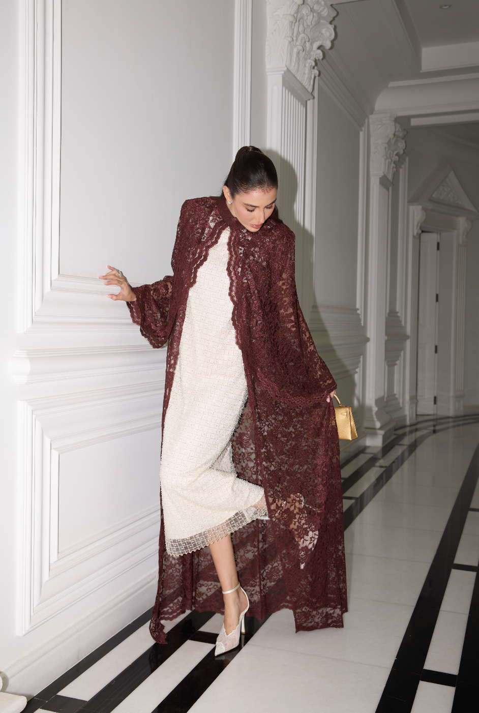 A woman poses in a hallway wearing a long, sheer burgundy lace kimono-style coat over a white textured dress. She is holding a small gold handbag and wearing white high heels.