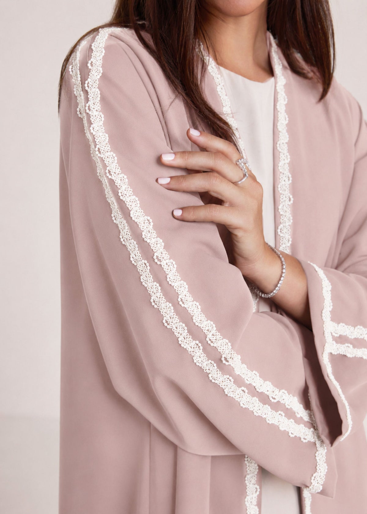 A close-up of a person wearing a dusty pink kimono-style cardigan with wide sleeves and white lace trim along the edges, over a white top.