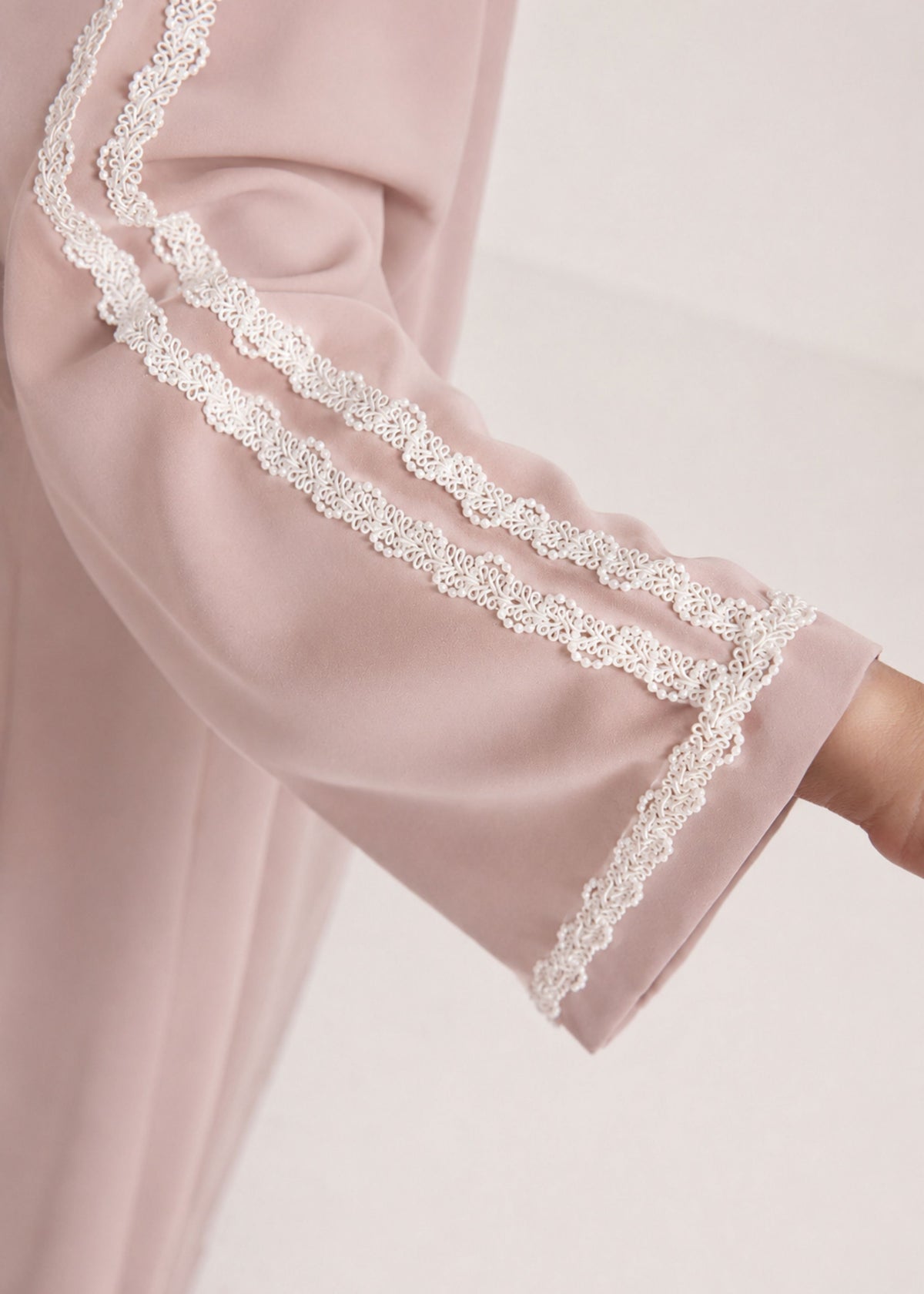 A close-up of a dusty pink garment sleeve featuring double decorative white lace trim along the cuff, worn by a model whose arm is visible.