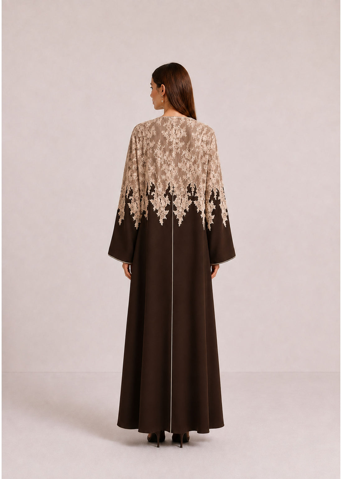 A woman stands against a plain light beige studio background wearing a long brown abaya dress with a beige floral lace overlay across the chest and upper sleeves, plus matching brown shoes.