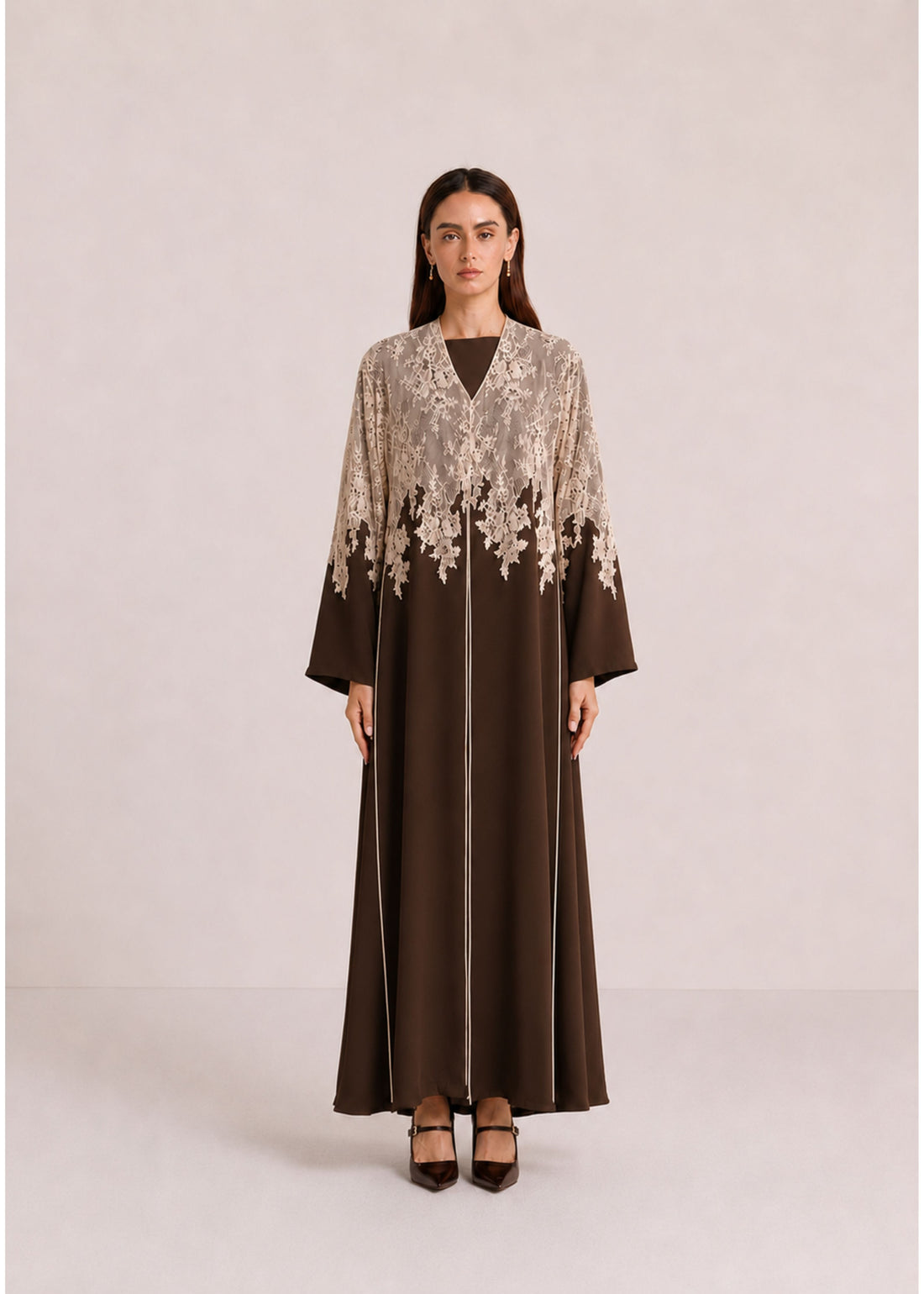 A woman stands against a plain light beige studio background wearing a long brown abaya dress with a beige floral lace overlay across the chest and upper sleeves, plus matching brown shoes.