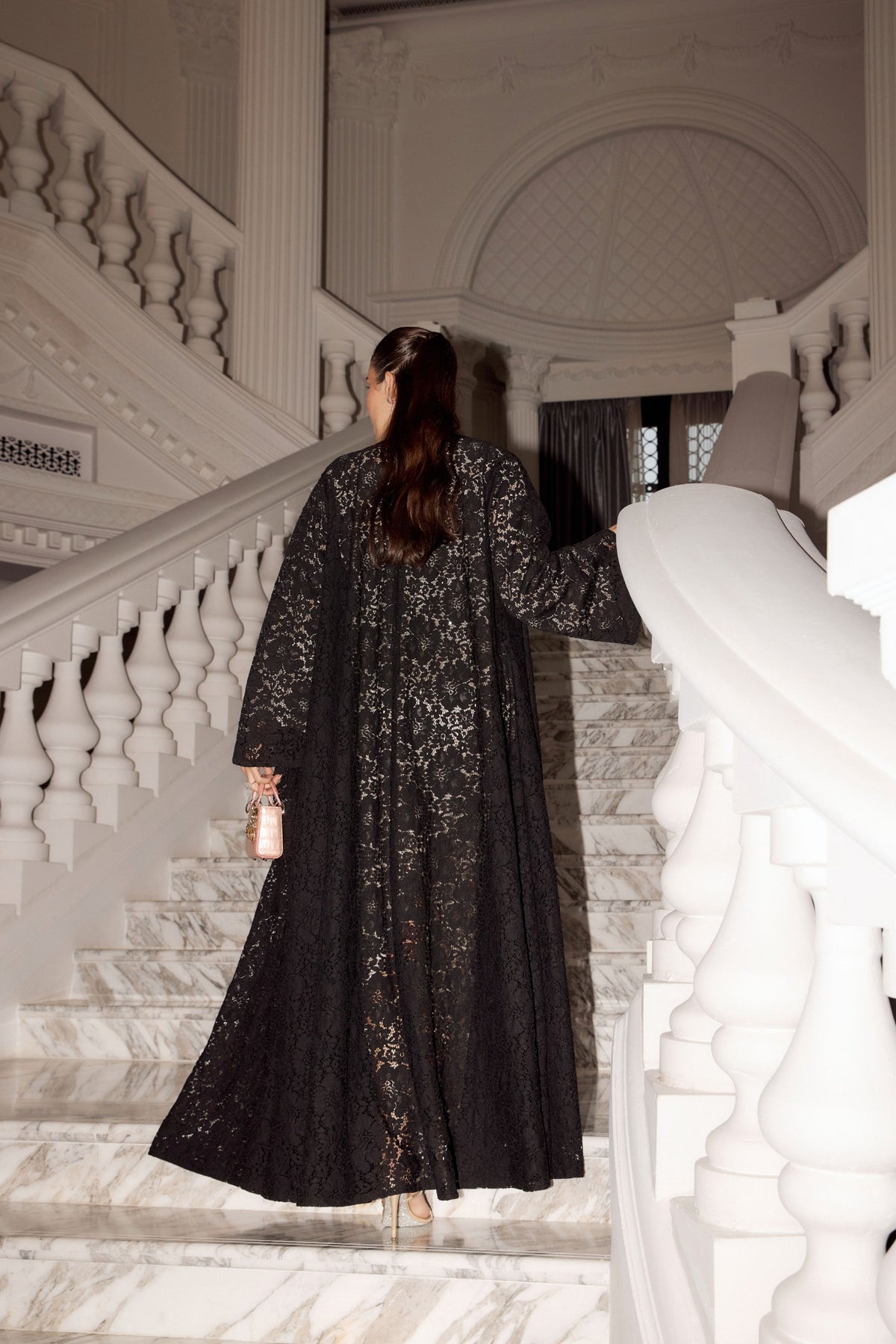 A woman wearing a long black lace abaya with gold floral embroidery is seen from behind as she walks down a grand white marble staircase in an elegant building.