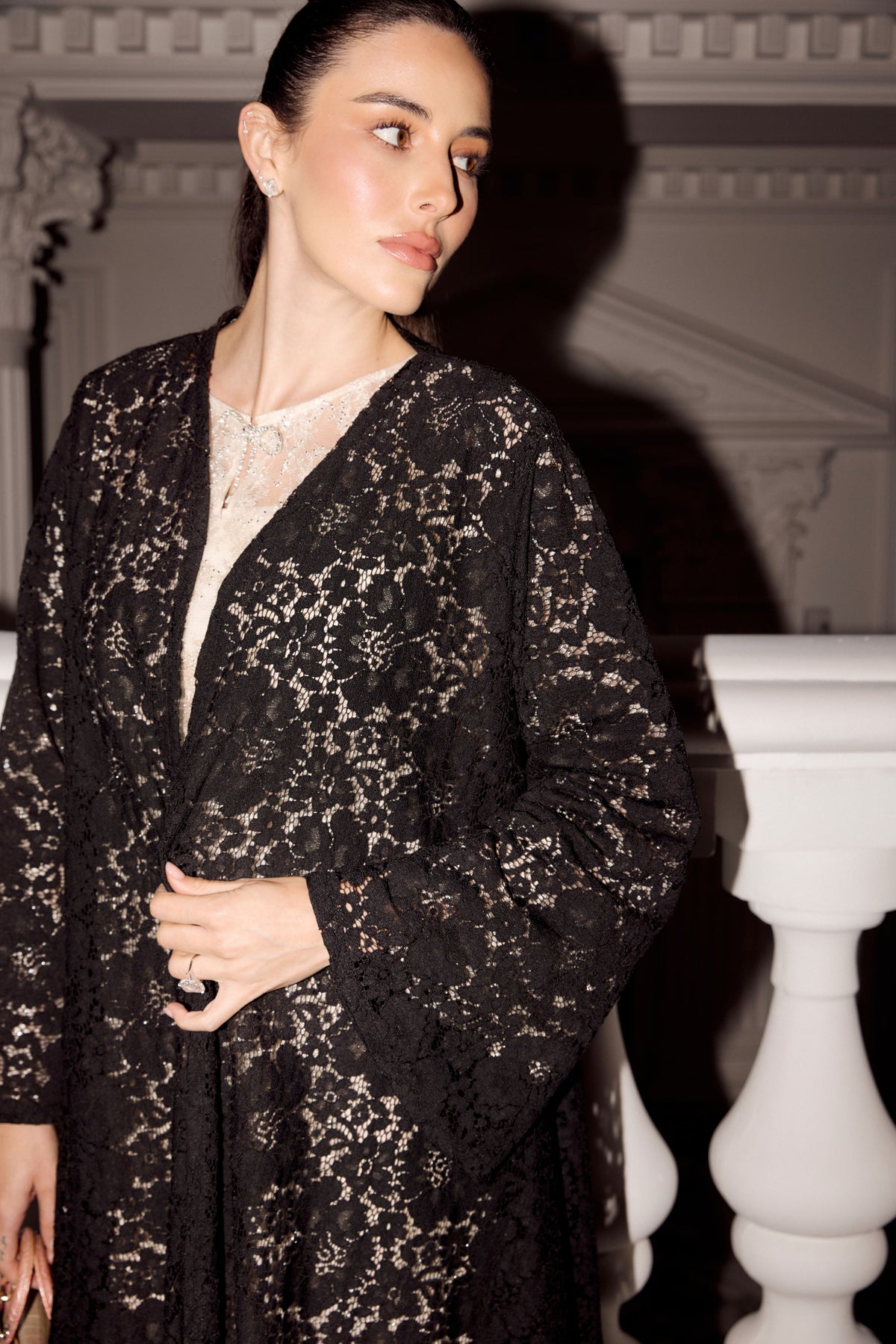A woman wearing a black lace cardigan over a cream lace top, standing in front of a white balustrade with a dark architectural background.