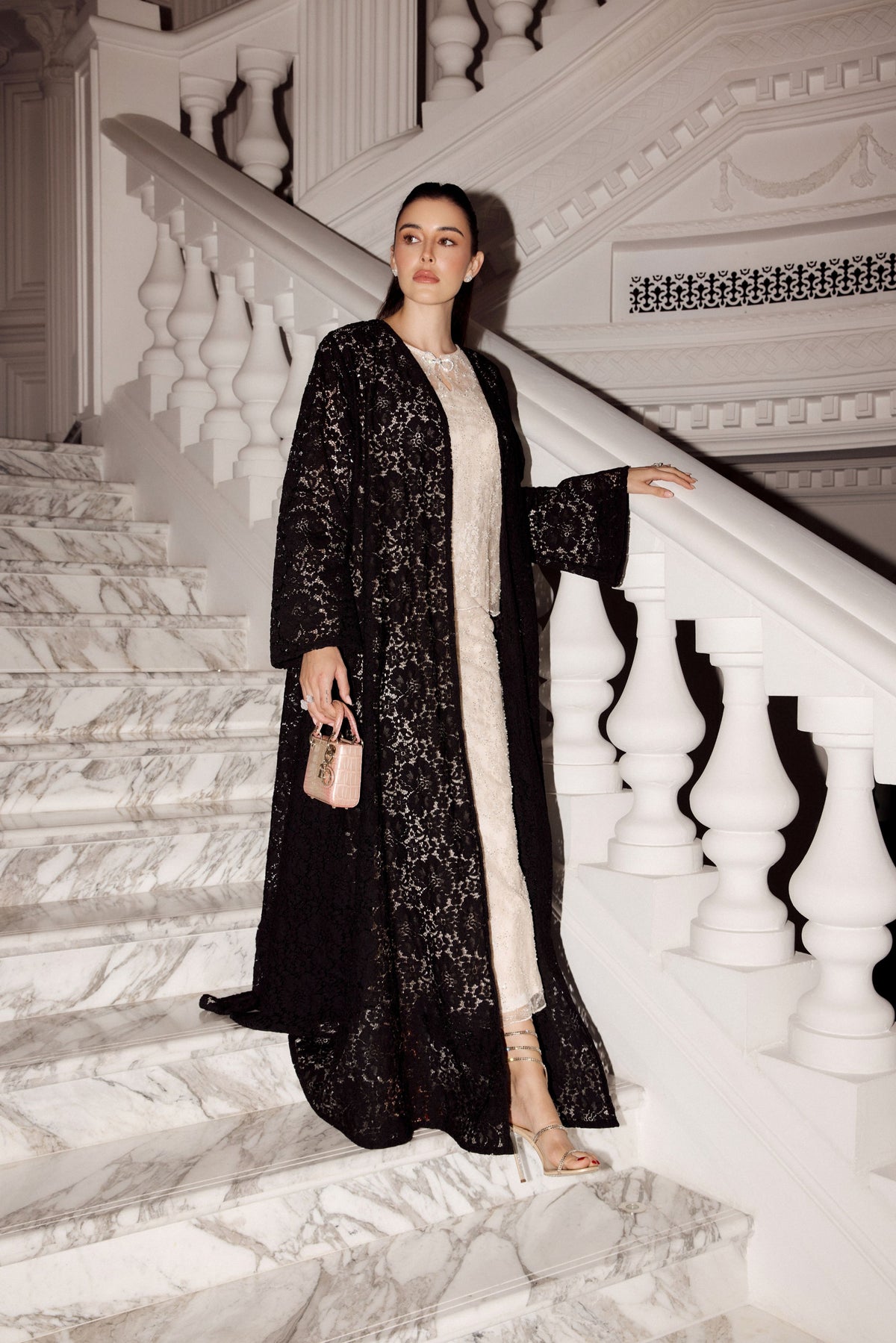A woman stands on white marble stairs wearing a long black embroidered abaya with a sheer black overlay, layered over a cream inner dress. She holds a small blush pink handbag.