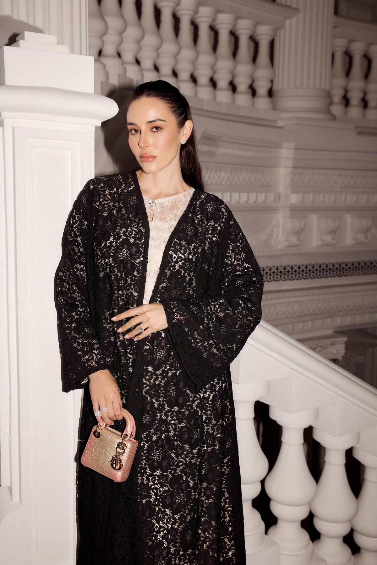 A woman stands in a grand white staircase hall wearing a black lace robe over a white top, holding a small pink handbag.